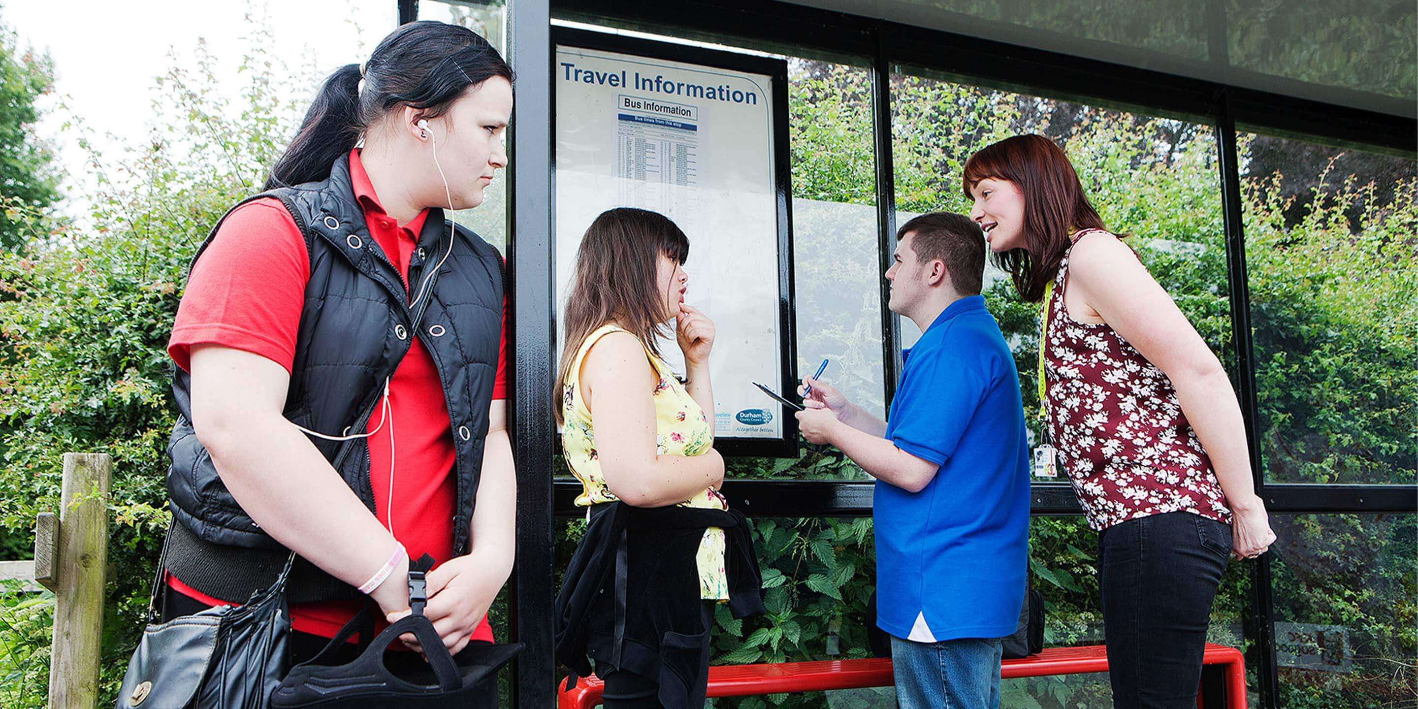 Group of young adults with a teacher at a bus stop looking at the bus timetable.