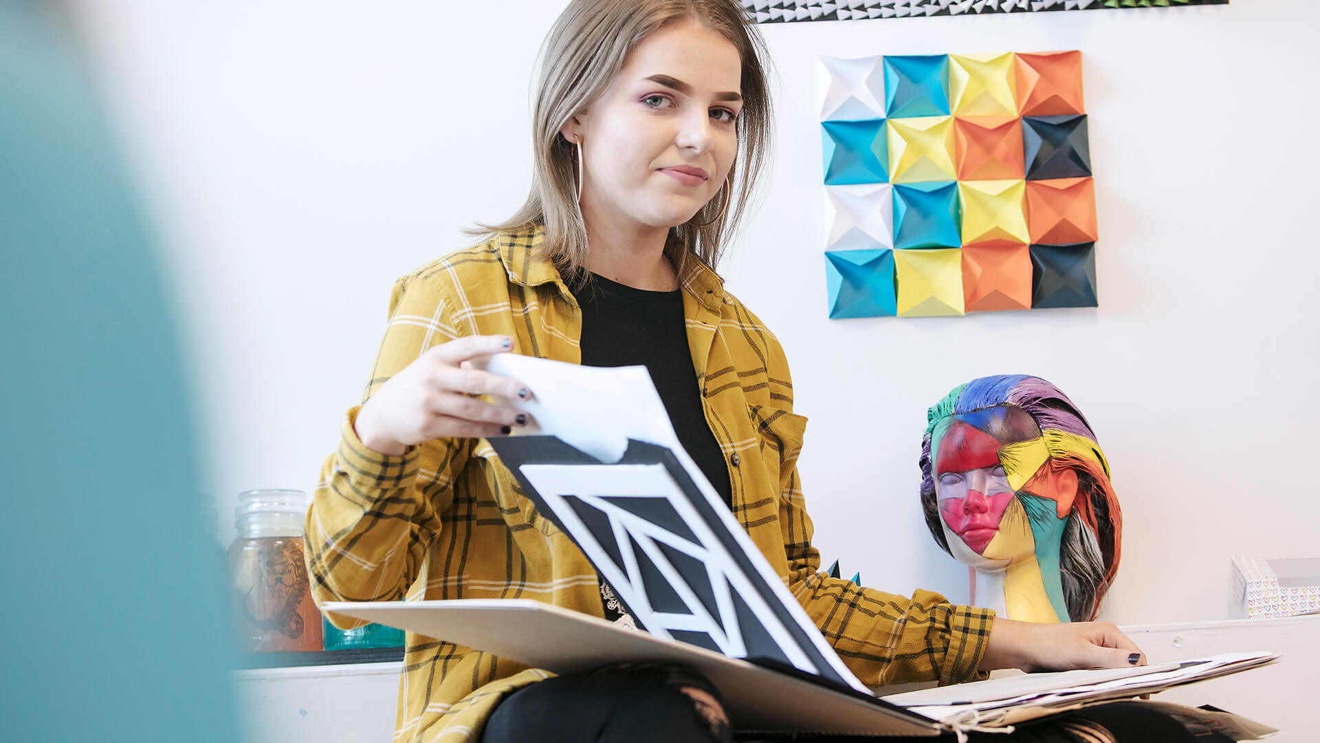 Young woman looking directly at the camera and smiling, whilst holding an art portfolio, with artwork behind her.