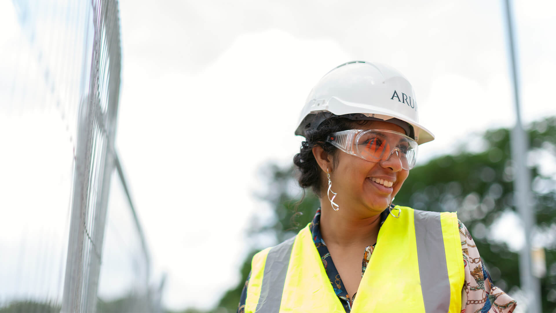 A woman wearing a white hard hat, high-vis jacket and safety goggles, smiling.