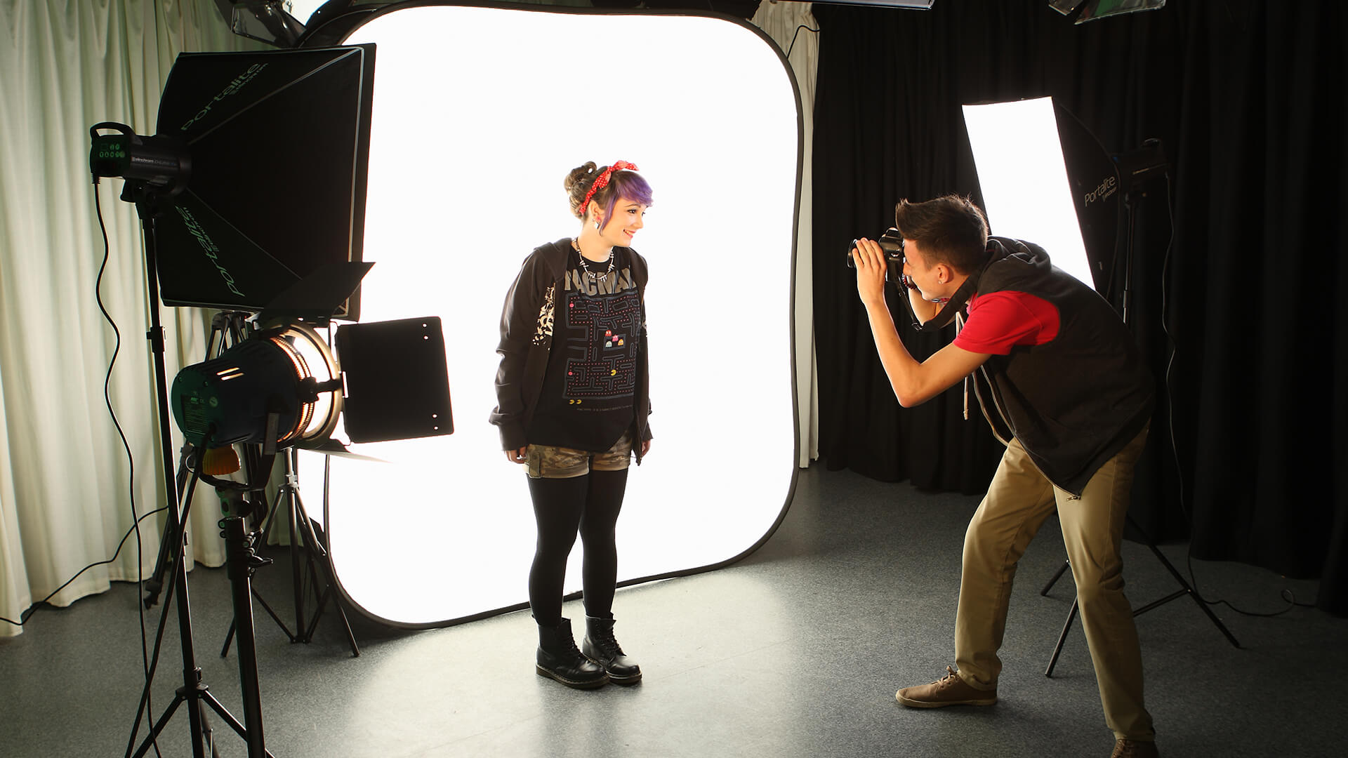 A young man taking pictures of a young woman that is standing in front of a light box.