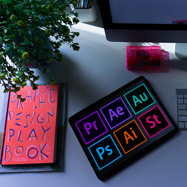 Desk with computer screen, keyboard, plant, graphic design book and smart tablet on it.