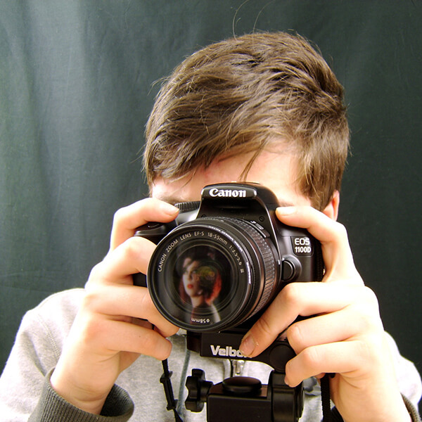 Young man holding a camera, taking a photograph. A young woman can be seen in the lens reflection.