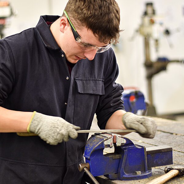 A young man wearing safety goggles and working with a piece of metal on a workbench.