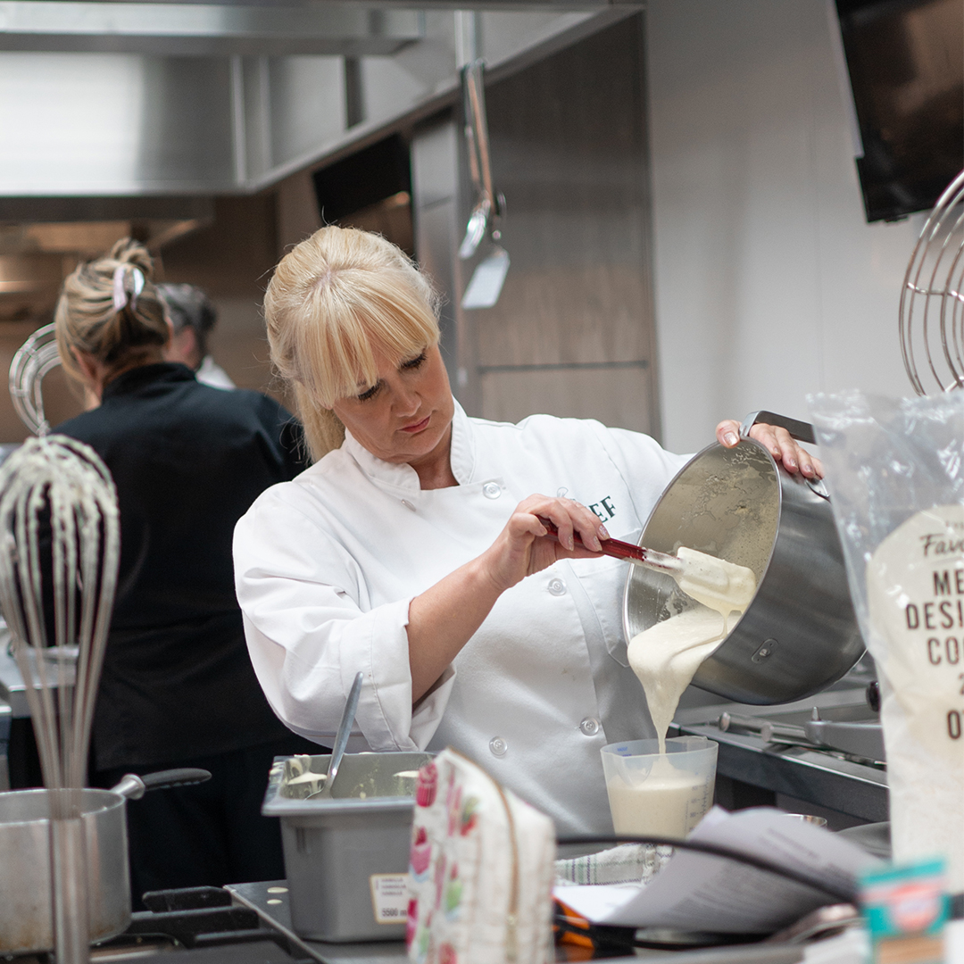 A middle aged women in chef's whites pouring a cream liquid from a mixing bowl into a jug