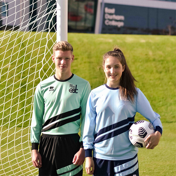 A young man and young woman standing of a football pitch next to a goal, wearing football uniforms and holding a football.