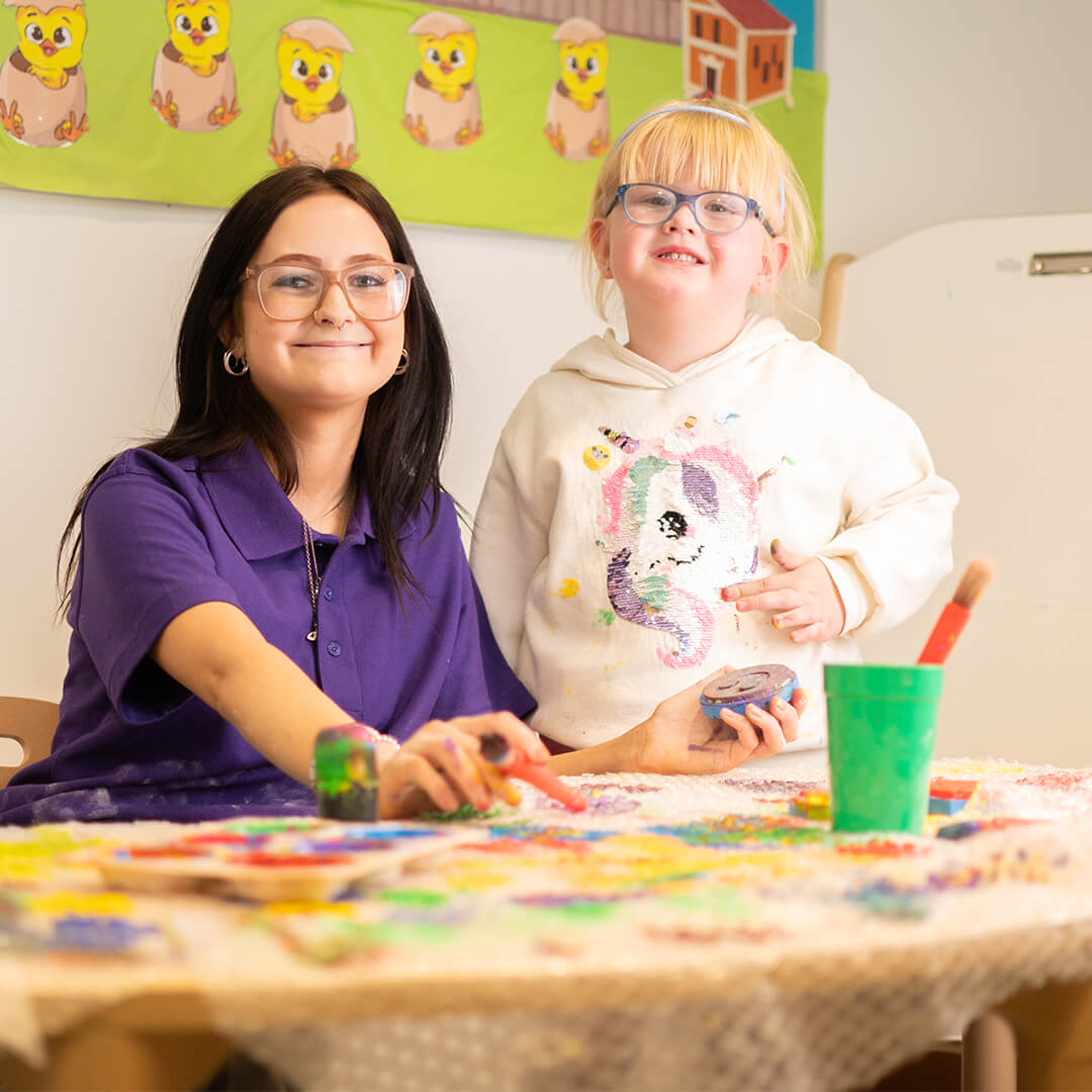 Smiling student in purple top with small child playing with paint
