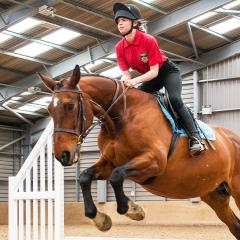 A student in a red top mid-air jumping on a horse over a jump