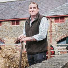 Photo of Karl Brown stood leaning on a spade next to some hay with stone buildings in the background