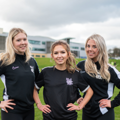 Three female football players standing on the college football pitch
