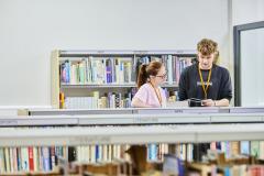 Two students talking in library behind shelf of books